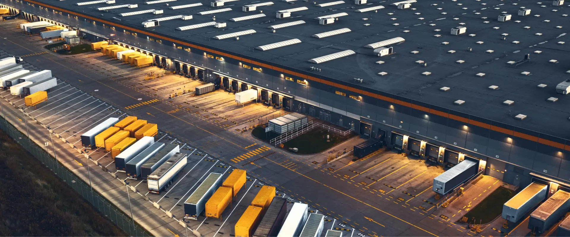 Luftaufnahme eines gro&szlig;en Distributionszentrums in der D&auml;mmerung mit Lkw-Anh&auml;ngern an den Laderampen sowie abgestellten Aufliegern und Wechselbr&uuml;cken.