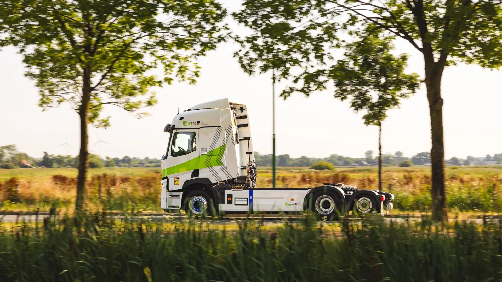 Witte elektrische ZEFES-truck rijdt zijwaarts over een landweg langs bomen en velden