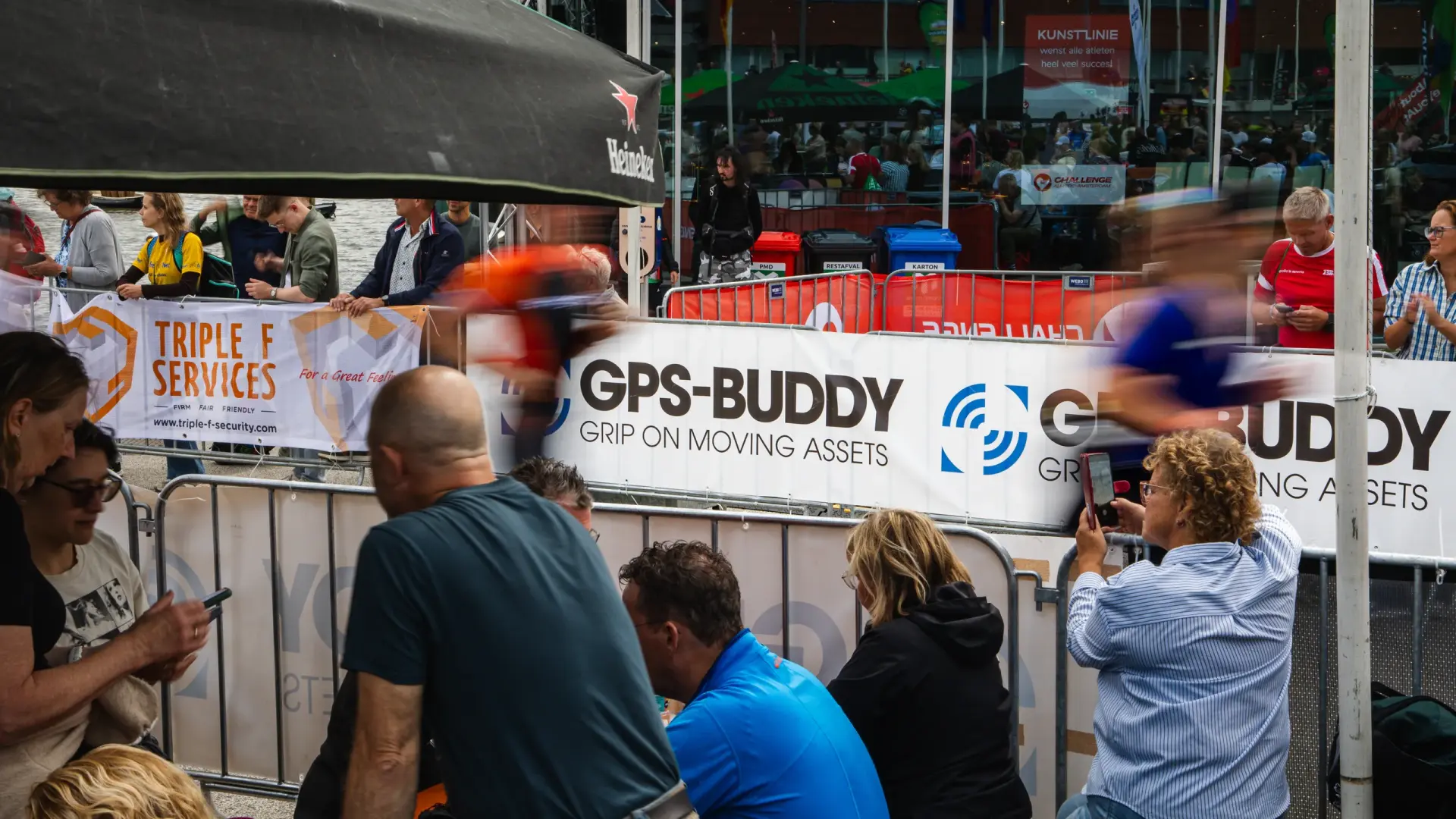 Zuschauer an der Strecke vor einem GPS-Buddy Banner, w&auml;hrend ein Triathlet verschwommen vorbeil&auml;uft