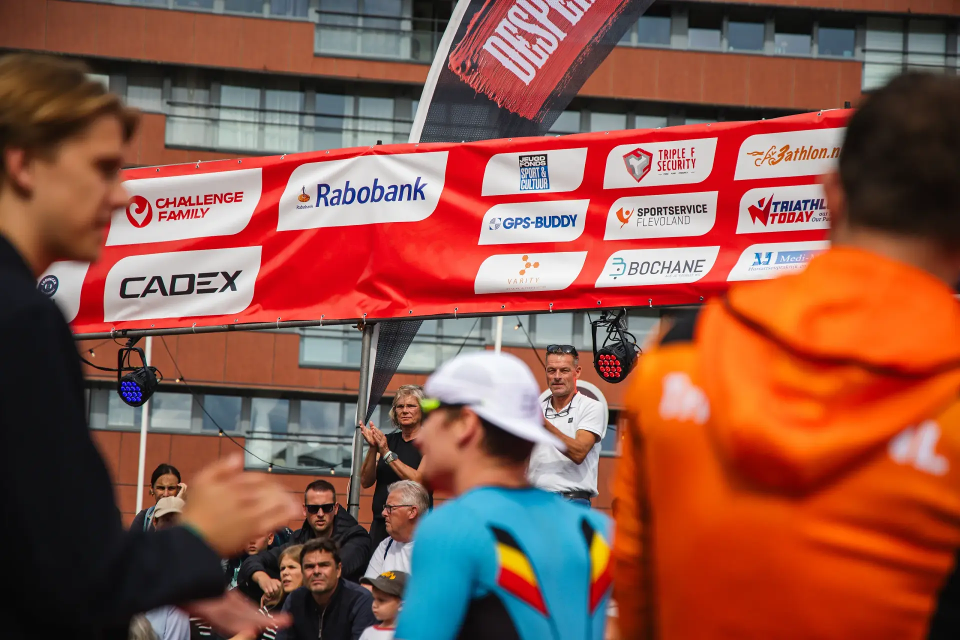 Sponsorenbanner mit Logos von u. a. Rabobank und GPS-Buddy &uuml;ber Zuschauern, im Vordergrund unscharfe Athleten