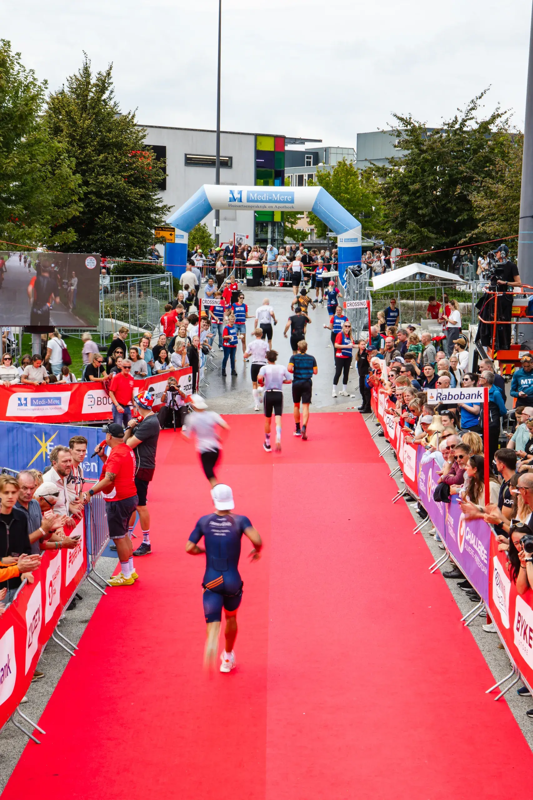 Triathleten laufen &uuml;ber einen roten Teppich zwischen Zuschauern, im Hintergrund ein aufblasbarer Bogen