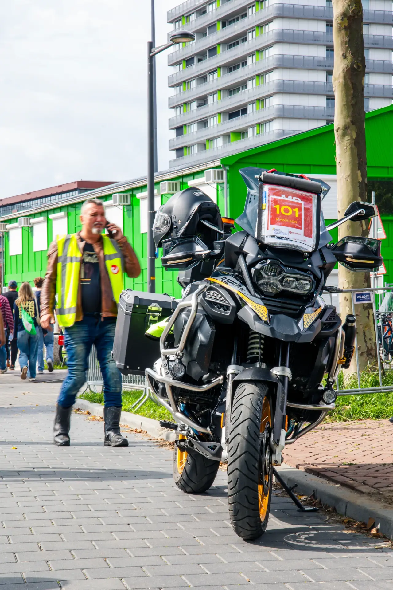 Mann in gelber Warnweste telefoniert neben einem schwarzen Motorrad mit Schild 101 Manager