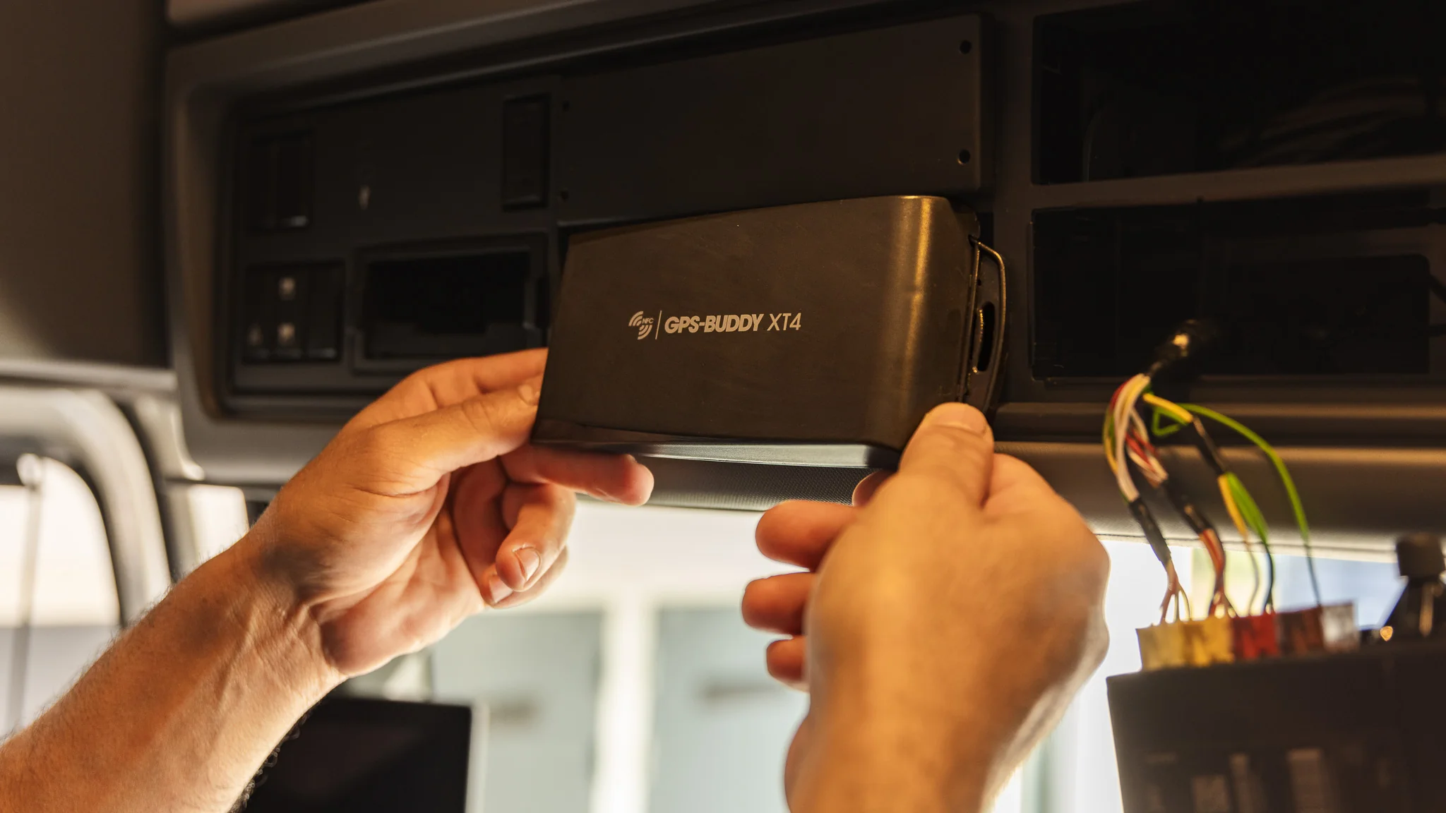 Close-up of hands holding a GPS-Buddy XT4 tracker at the overhead bay, wiring alongside
