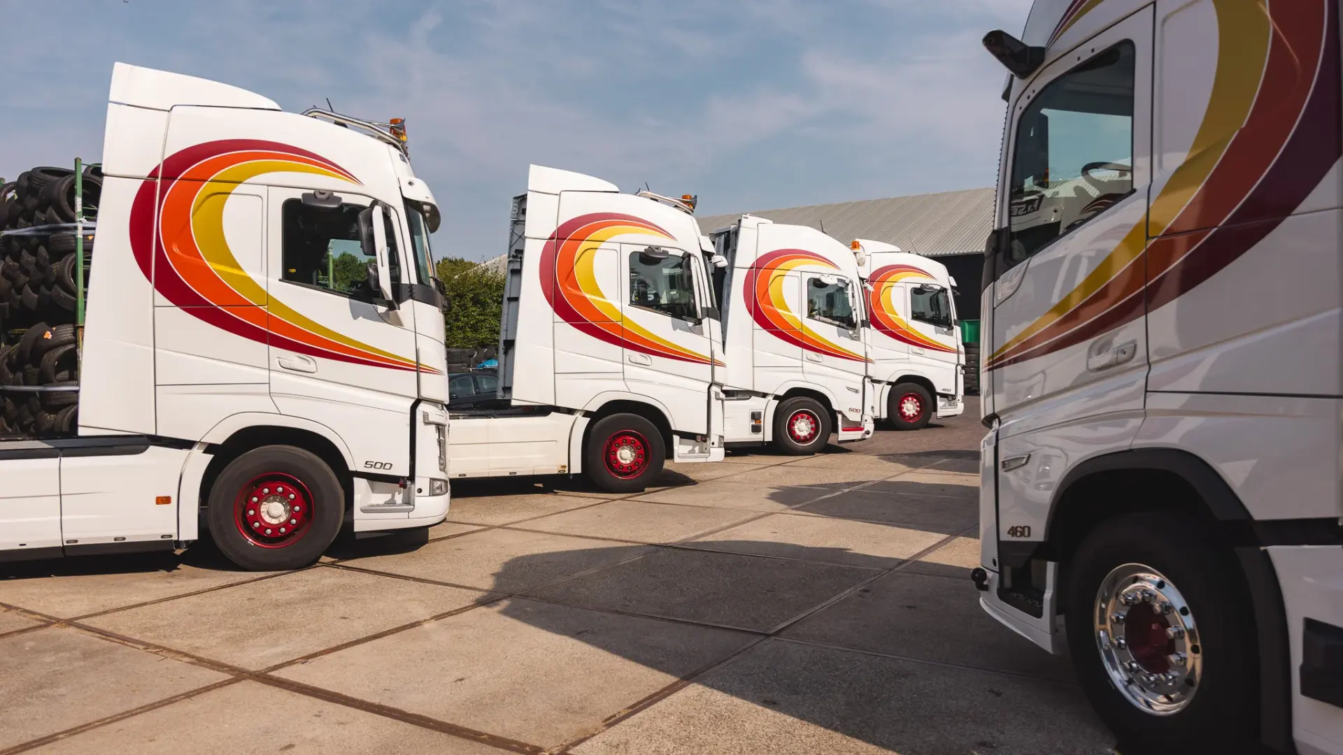 Rij witte Volvo-trucks met rood-oranje-gele sierstrepen, schuin geparkeerd op een geplaveid terrein